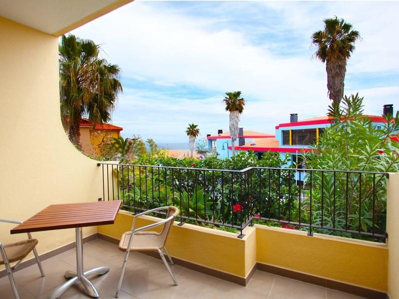Balcone con sedie e tavolo, vista su palme e case sotto un cielo azzurro.