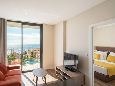 Helles Wohnzimmer mit Meerblick, rotem Sofa, kleinem Tisch und angrenzendem Schlafzimmer mit gelber Bettdecke.