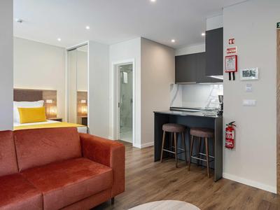 Moderner Wohnraum mit rotem Sofa, kleiner Küchenzeile, Barhockern und Holzboden in neutralen Farben.