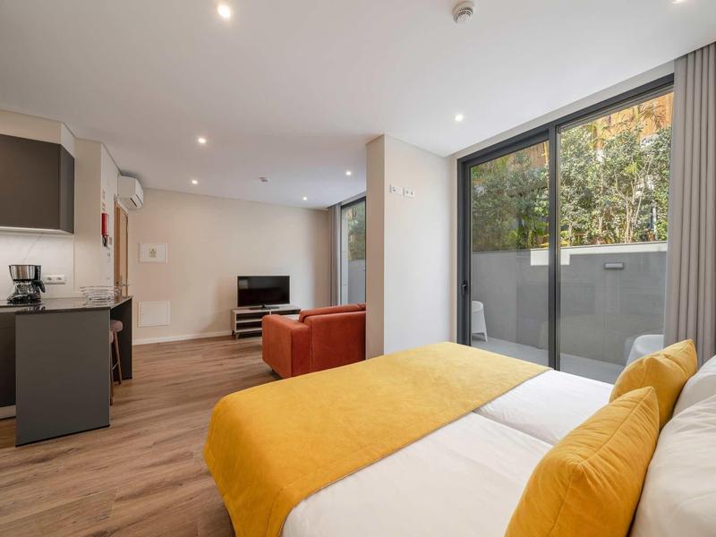 Moderner Wohnraum mit Bett, orange-gelben Decken, Fernseher, Küchenzeile und großem Fenster.