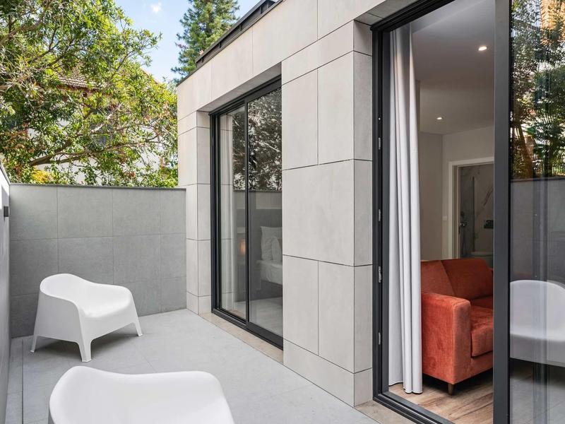 Moderner Balkon mit grauen Fliesen, weißen Stühlen und Blick auf grüne Bäume im Hintergrund.