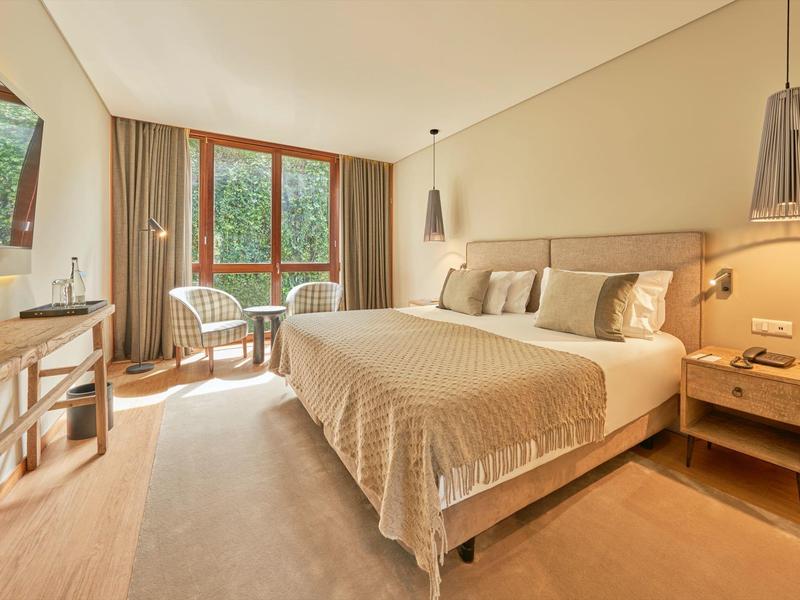 Camera d'hotel luminosa con letto matrimoniale, mobili in legno e due sedie vicino alla finestra con vista sul verde.