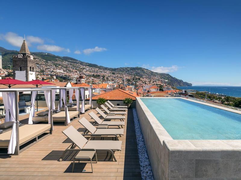 Infinity zwembad met ligstoelen met uitzicht op een kustplaats onder een blauwe lucht.