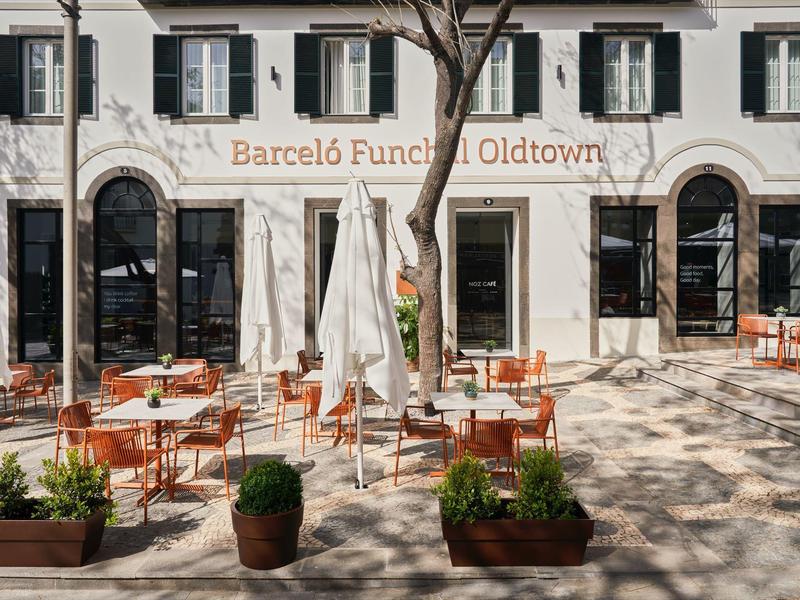 Buiten zitgedeelte met tafels, stoelen en bomen voor een wit hotelgebouw.