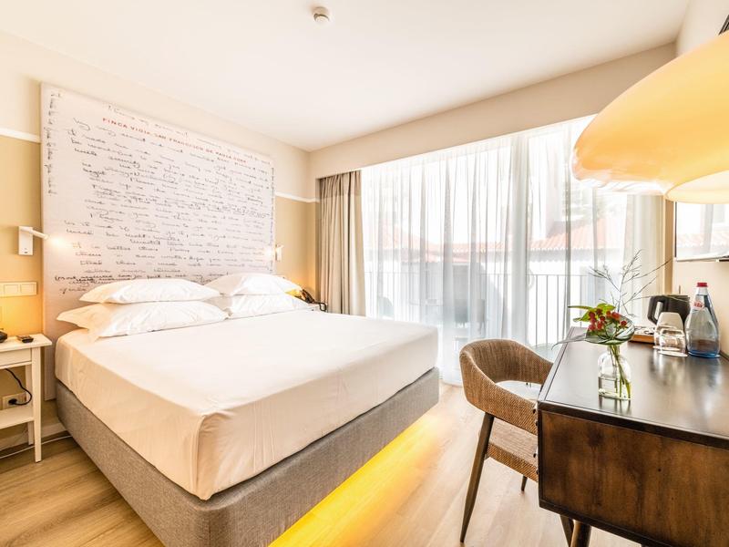 Chambre d'hôtel lumineuse avec lit double, bureau, chaise et balcon avec rideaux.