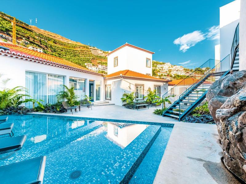 Espace piscine moderne avec chaises longues, bâtiment d'hôtel adjacent et arrière-plan montagneux sous le soleil.