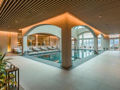 Modern hotel indoor pool with loungers, large windows, and warm lighting.