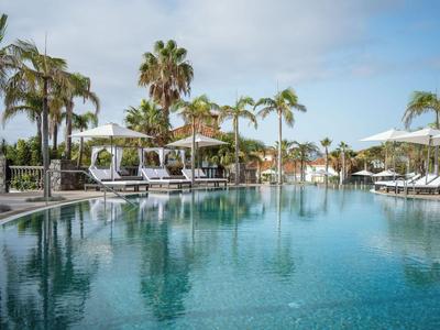 Großer Außenpool mit Liegestühlen und Palmen in einer Hotelanlage bei klarem Himmel.