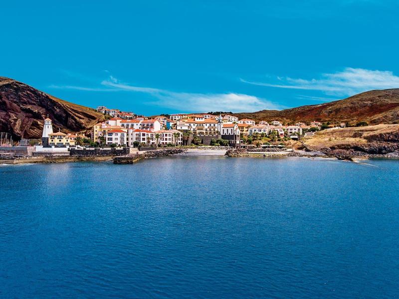 Small coastal village with white houses by calm blue bay under clear sky.