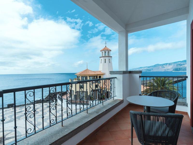 Balcony with table and chair overlooking lighthouse and sea under blue sky.