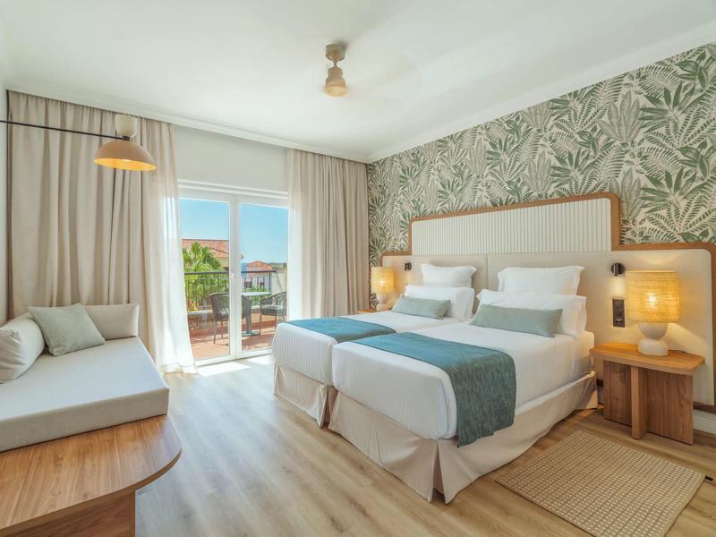 Bright hotel room with double bed, sofa, palm-patterned wall, and balcony overlooking the sea.