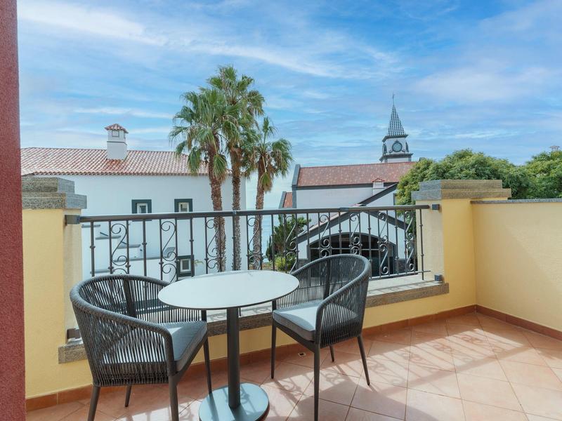 Balcony with two chairs and small table overlooking palm trees and white buildings.