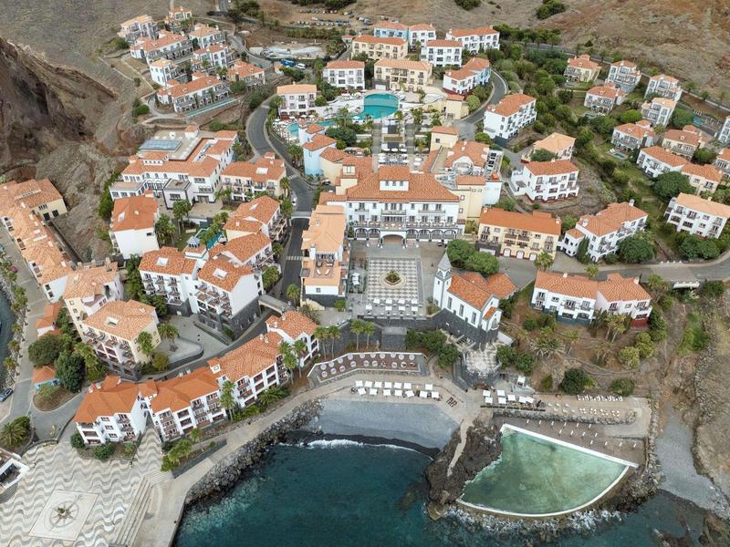 Aerial view of a resort with red roofs, pool, beach, and sea view in a rocky coastal landscape.