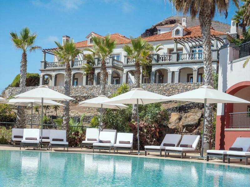 Hotel with palm trees and pool under blue sky on a sunny day.