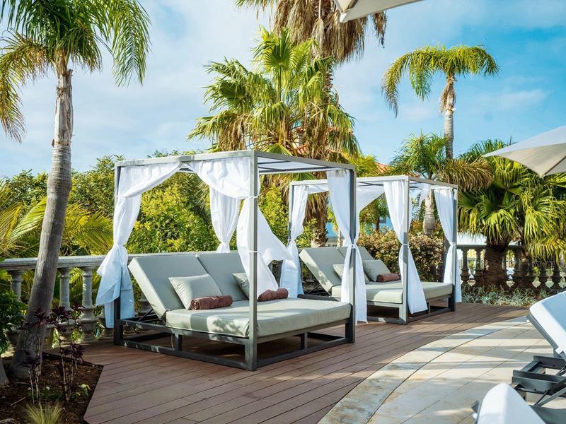 Several comfortable daybeds with white curtains on a terrace with palm trees.