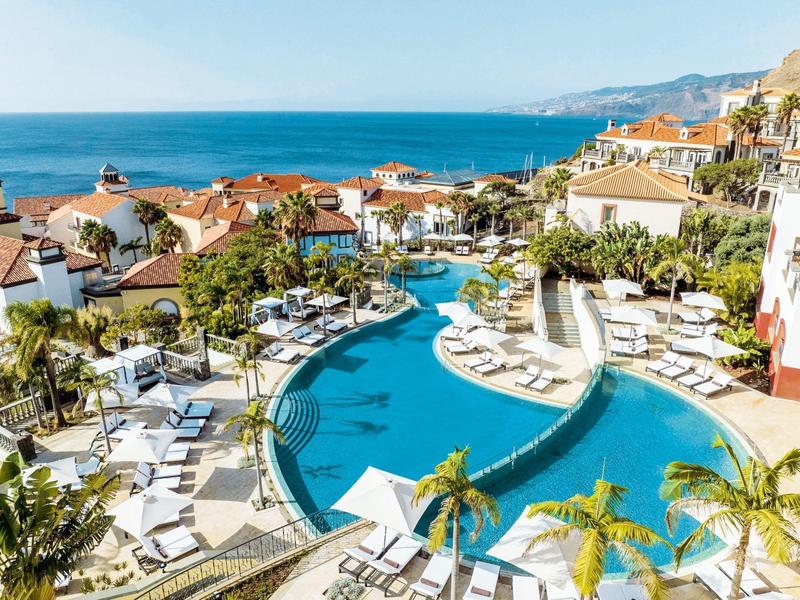 View of a pool with sun loungers and palm trees next to white houses and sea view.