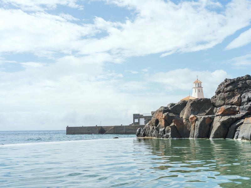 View of a rocky pool with sea and lighthouse in the background under a cloudy sky.