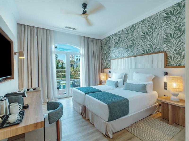 Bright hotel room with two single beds, plant-patterned wall, and balcony view.