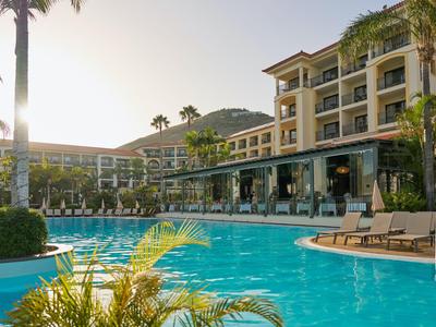 Complejo hotelero moderno con piscina grande, tumbonas y palmeras bajo luz cálida.
