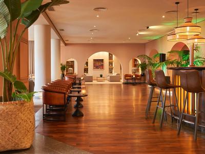 Un vestíbulo de hotel cálido con suelo de madera, sillones marrones, taburetes de bar y plantas verdes.