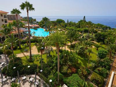 Complejo hotelero con piscina, jardines exuberantes y vista al mar en un día despejado.