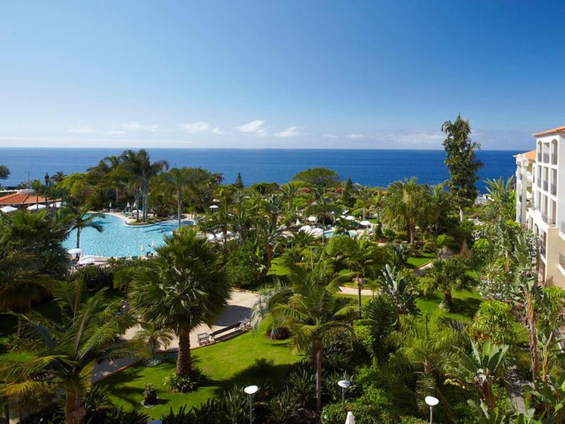 Blick auf tropischen Hotelgarten mit Pool, Palmen und Meer im Hintergrund unter klarem Himmel.