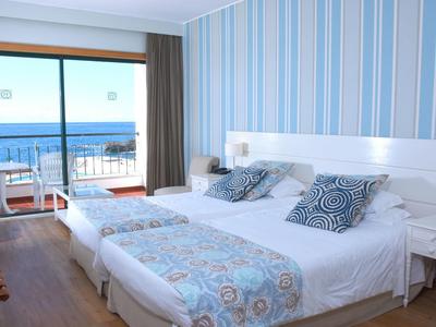 Schlafzimmer mit zwei Einzelbetten, blau-weiß gestreifter Wand, Balkontür mit Meerblick und Balkonmöbeln.