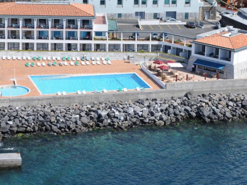 Hotelanlage mit rechteckigem Außenpool, Liegestühlen und einer Hafenmauer aus grauen Steinen am Wasser.