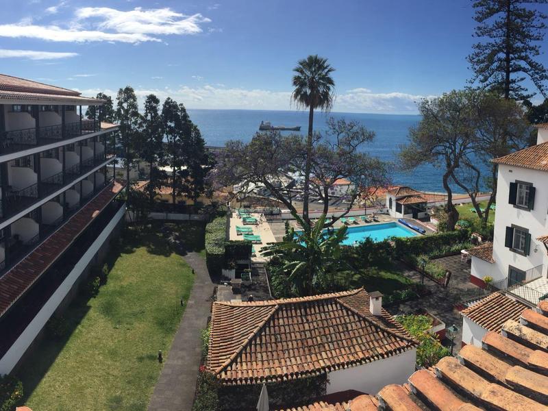 Blick auf Häuser, Pool, Bäume und Meer unter blauem Himmel mit vereinzelten Wolken.