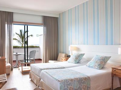 Stanza d'albergo con due letti singoli, scrivania, sedie e balcone con vista su palme.