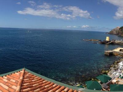 Vista panoramica di una città costiera con mare, piscina e tetti rossi sotto un cielo azzurro.