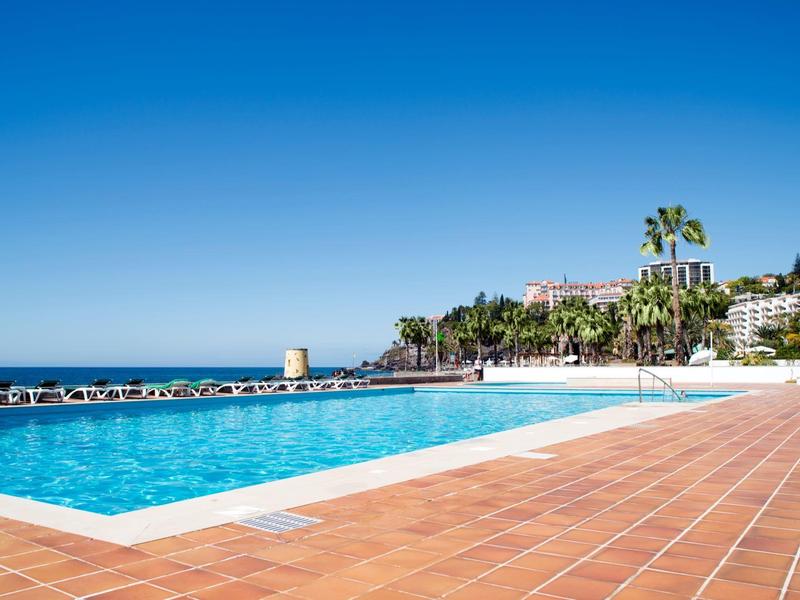 Piscina azzurra limpida con piastrelle terracotta e palme sotto un cielo azzurro.