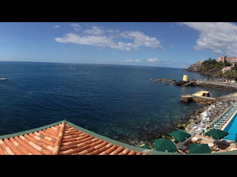 Vista panoramica di una città costiera con mare, piscina e tetti rossi sotto un cielo azzurro.