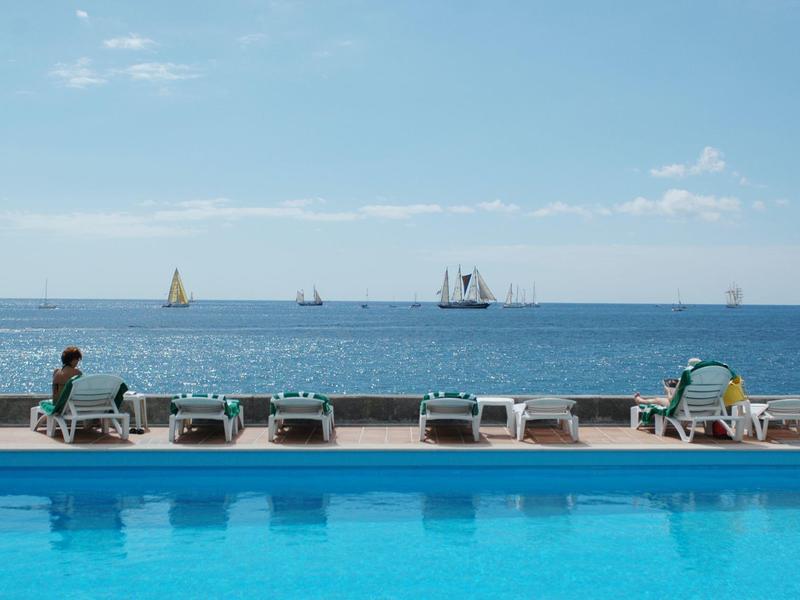 Piscina blu con lettini e persone, barche a vela sul mare calmo sotto cielo sereno.