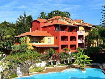 Hotel su più piani in colori caldi con piscina, circondato da piante tropicali e cielo azzurro.