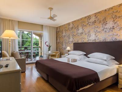 Camera d'hotel luminosa con letto matrimoniale, carta da parati floreale e balcone con vista sul verde.