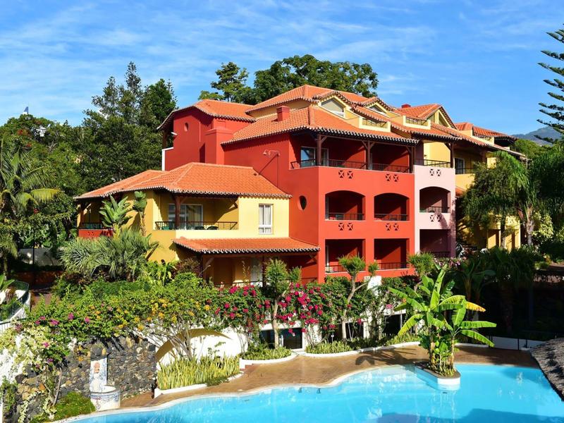 Hotel su più piani in colori caldi con piscina, circondato da piante tropicali e cielo azzurro.