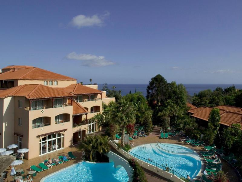 Complesso alberghiero con più piscine, circondato da palme e vista sul mare sotto un cielo azzurro.