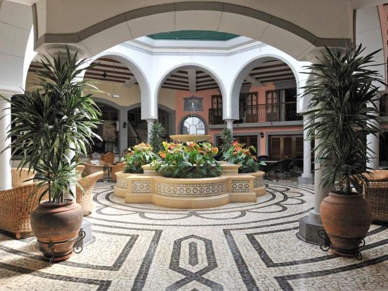 Cortile luminoso con fontana, grandi piante e galleria circostante in tonalità calde.