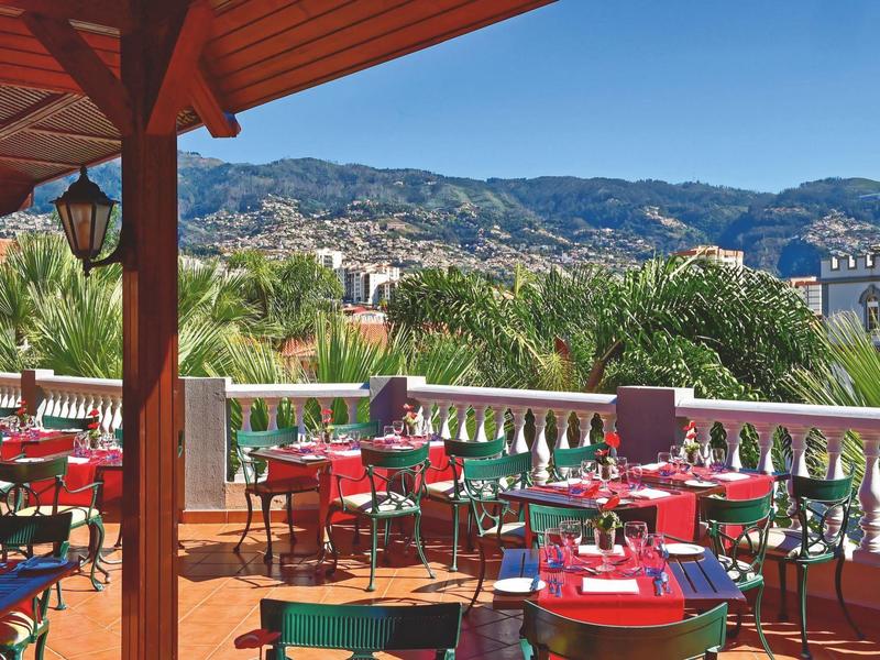 Terrazza con tavoli e sedie colorate, vista su palme e montagne con bel tempo.