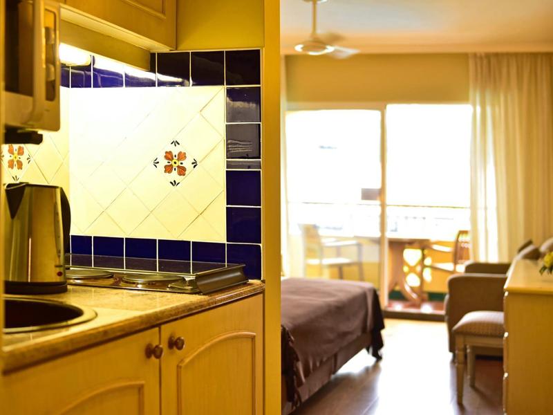 Camera d'hotel luminosa con letto, balcone e angolo cottura con piastrelle blu.