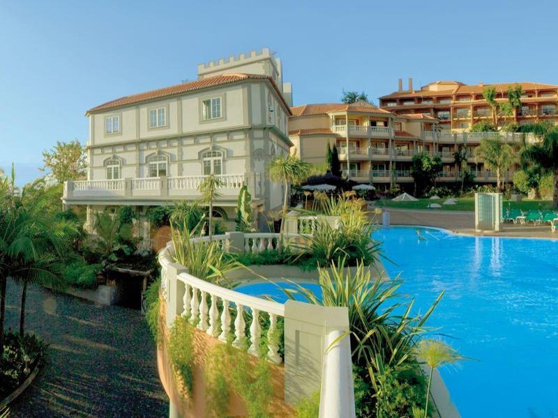 Complesso alberghiero con grande piscina, piante fiorite e cielo limpido.