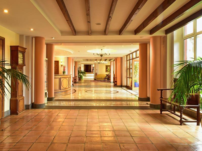 Ampio corridoio d'hotel con pavimento in terracotta, colonne e piante ai lati.