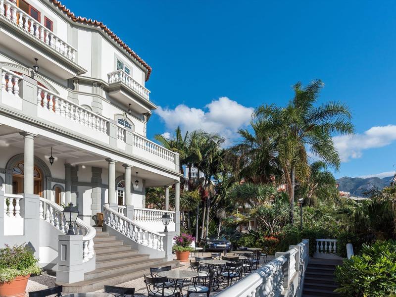 Grande edificio bianco dell'hotel con terrazza e tavoli all'aperto, circondato da palme.