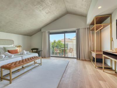 Jasny pokój hotelowy z dużym łóżkiem, drewnianą podłogą i balkonem z widokiem na drzewa.