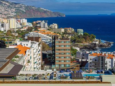 Vista de un hotel costero con piscina, rodeado de edificios y el mar al fondo.
