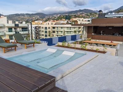 Piscina moderna de hotel con tumbonas y bar en la terraza, rodeada de edificios y montañas.