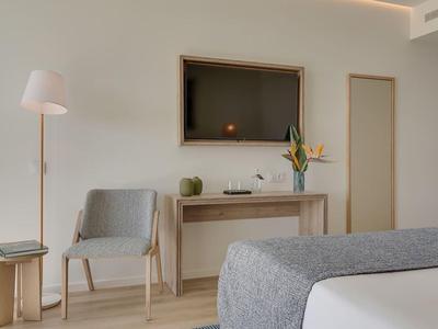 Jasny pokój hotelowy z szarym łóżkiem, krzesłem, drewnianą konsolą i dużym lustrem.
