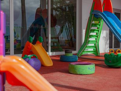 Bunter Kinderspielplatz mit Rutschen und Klettergeräten auf rotem Bodenbelag, Sonne scheint.