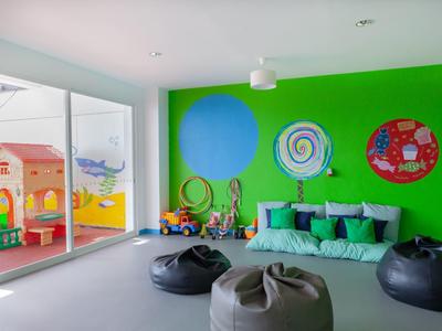 Kinderspielzimmer mit grüner Wand, bunten Dekorationen, Sofa und Sitzsäcken vor großen Fenstern.